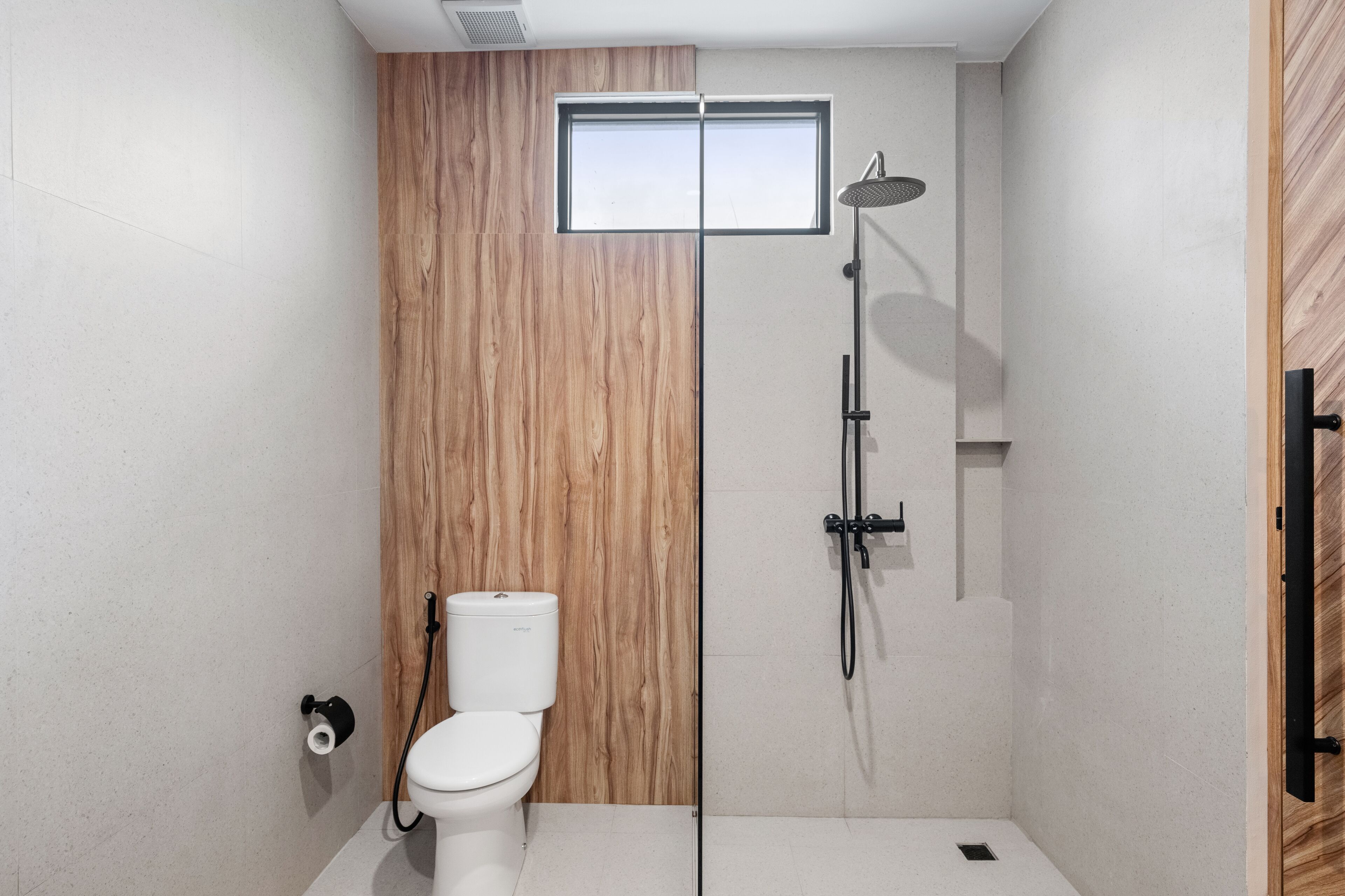 A modern bathroom with a wooden panel wall, a white toilet, a shower with a handheld showerhead, and a small window above the toilet.
