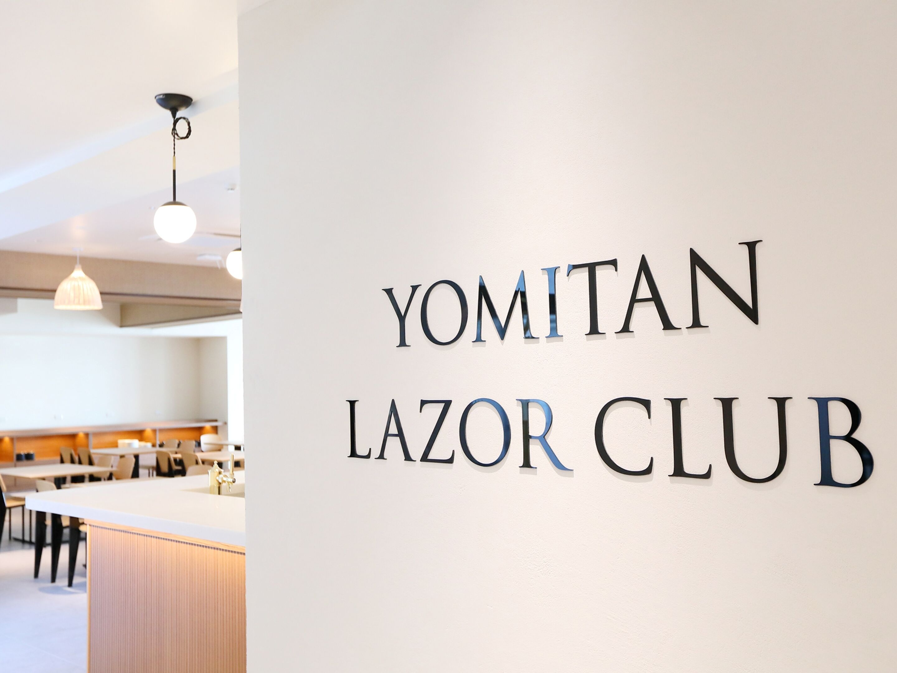 Ein Schild mit der Aufschrift 'YOMITAN LAZOR CLUB' in einem modernen Innenraum mit weißer Wand und hängenden Pendelleuchten.