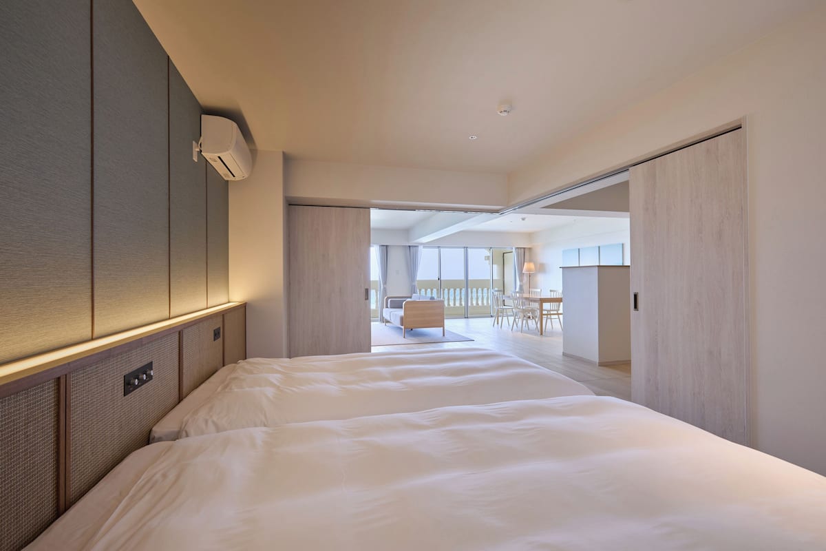 A modern hotel room with a large bed, a ceiling-mounted air conditioner, and a view of the adjacent dining area.