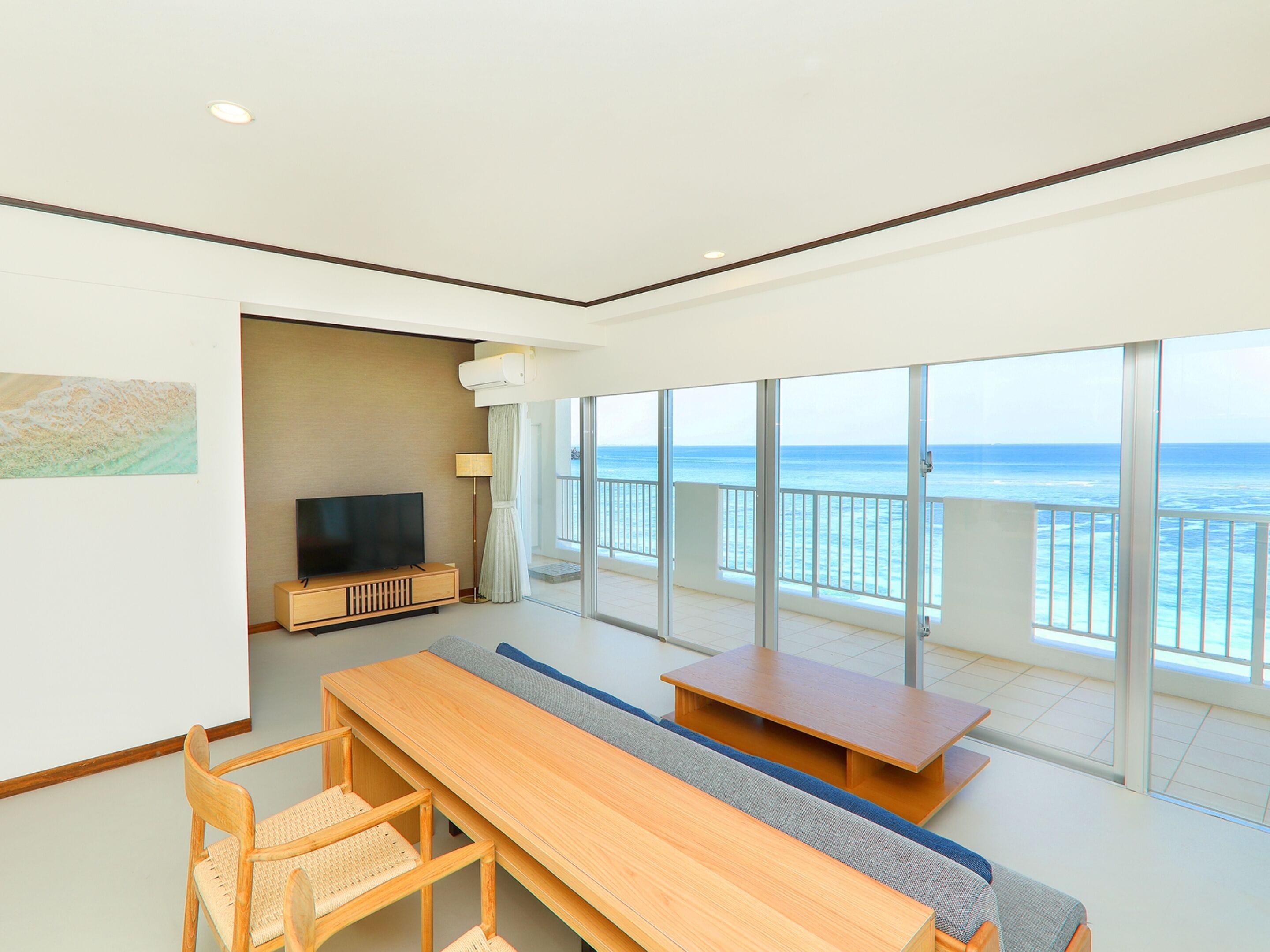A modern hotel room with a balcony, a flat-screen TV, a wooden dining table, and a sofa.