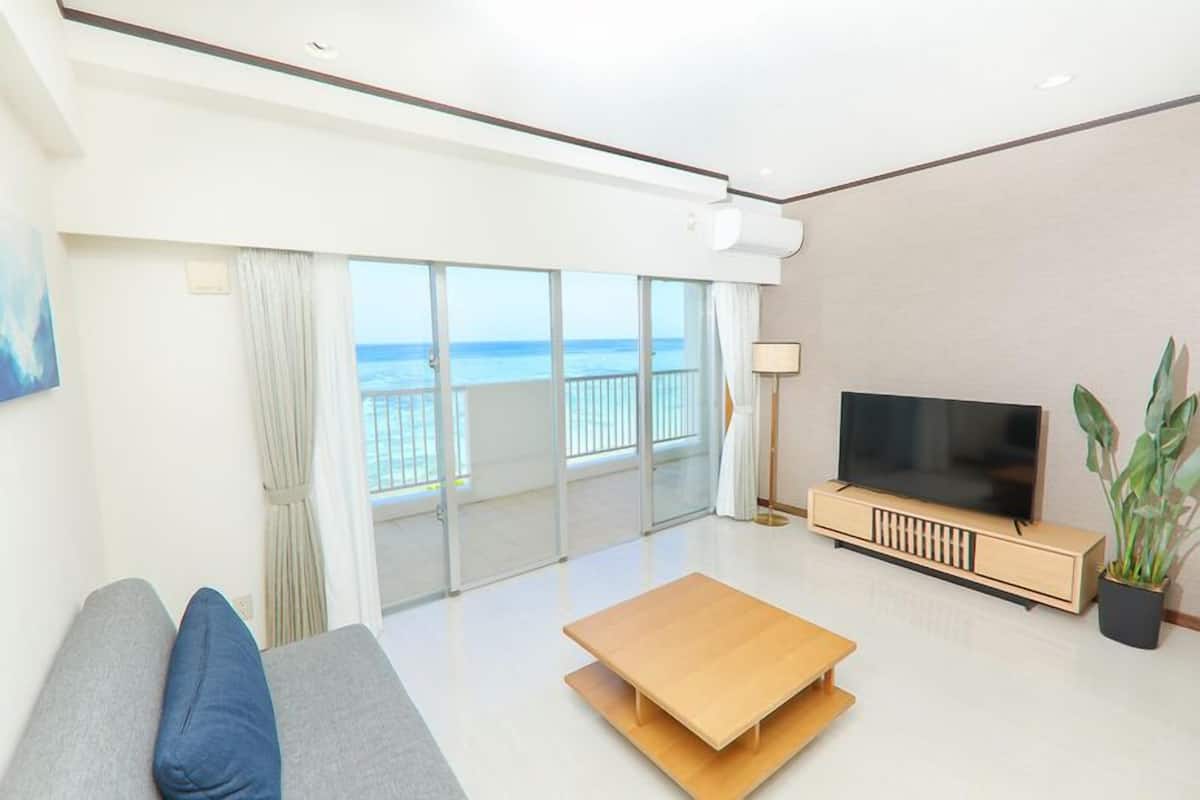 A modern living room with a sofa, a coffee table, a TV, and a balcony with a view of the sea.