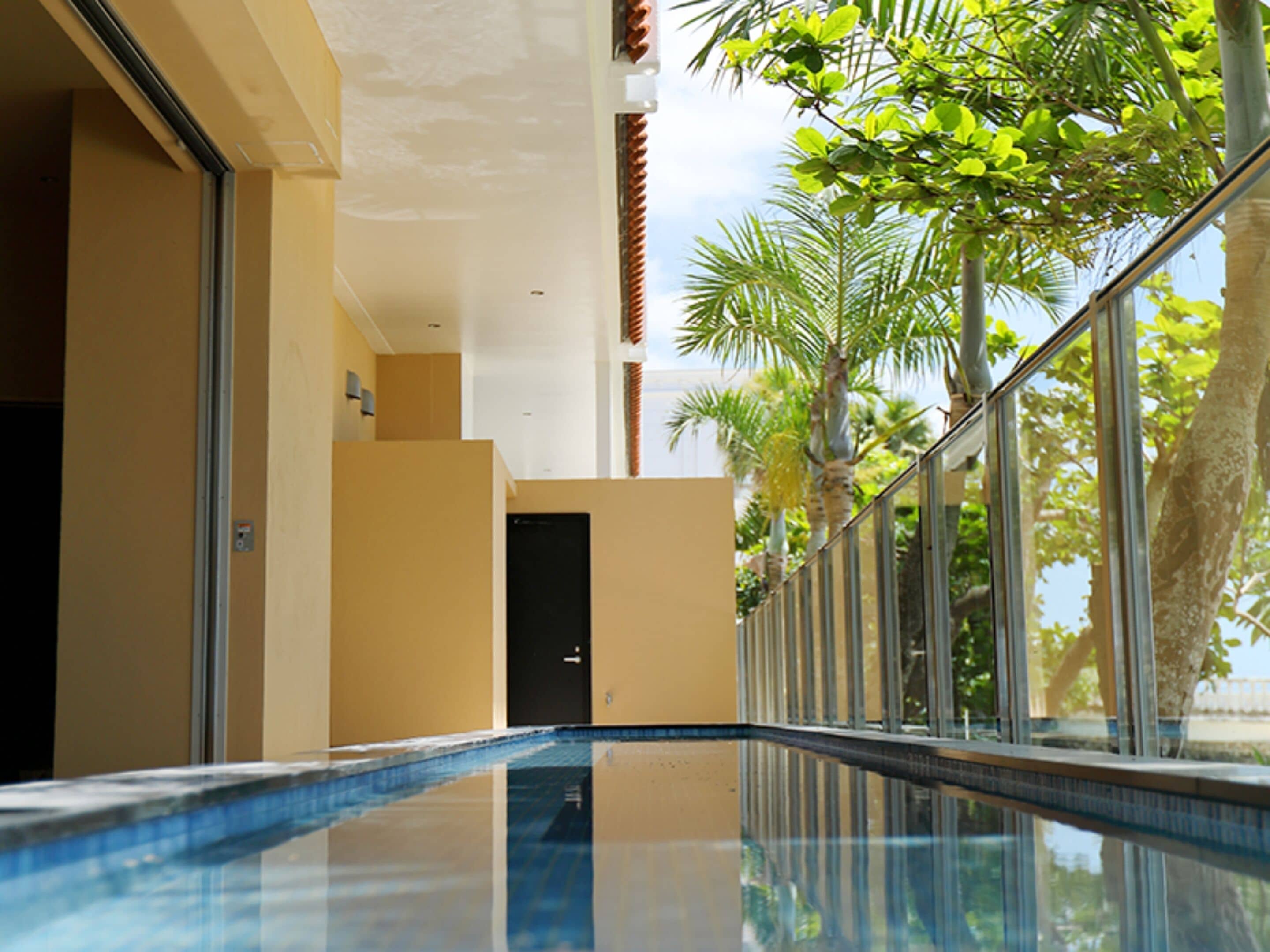 A swimming pool with a glass barrier, a door, and a view of palm trees and a building.