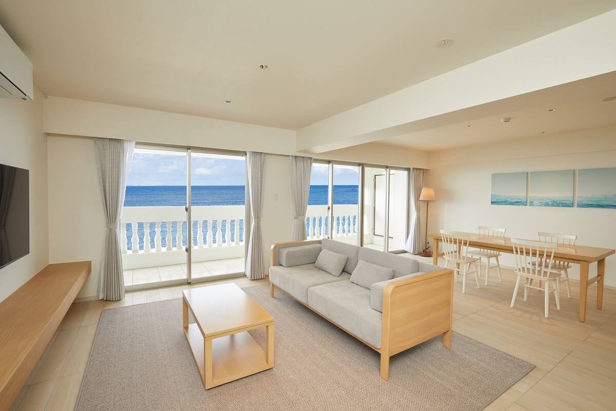 A spacious living room with a large sofa, a coffee table, and a dining area with chairs and a table. The room has large windows with a view of the sea.
