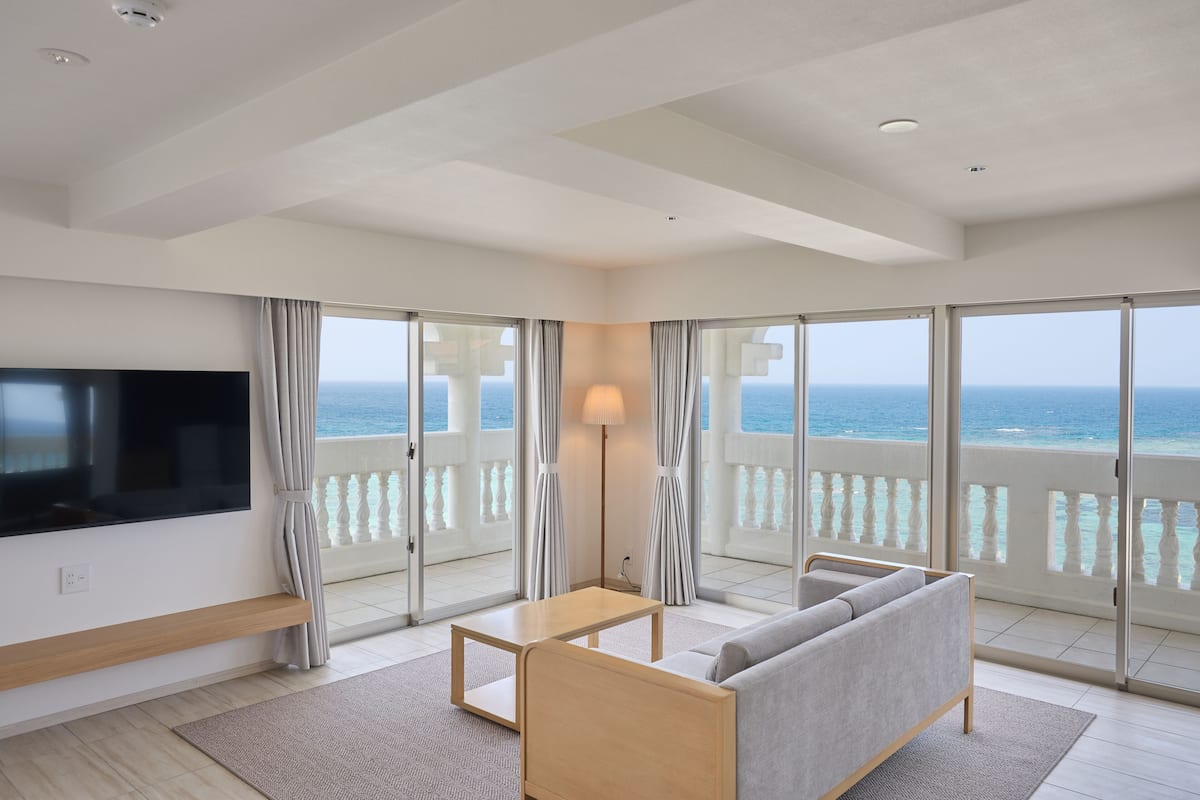 A modern living room with a flat-screen TV, a wooden coffee table, a grey sofa, and a lamp. Large windows offer a view of the ocean.