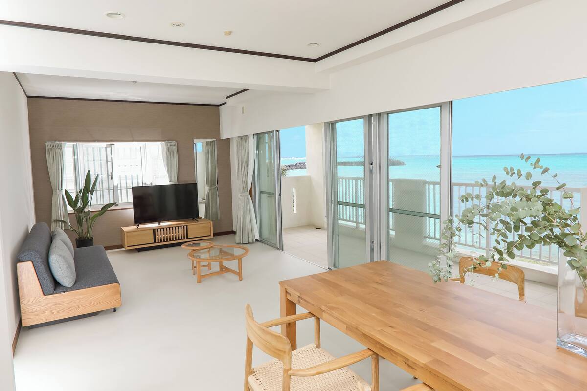 A modern living room with a sofa, TV, wooden table, and chairs. It features large glass doors leading to a balcony with a view of the sea.