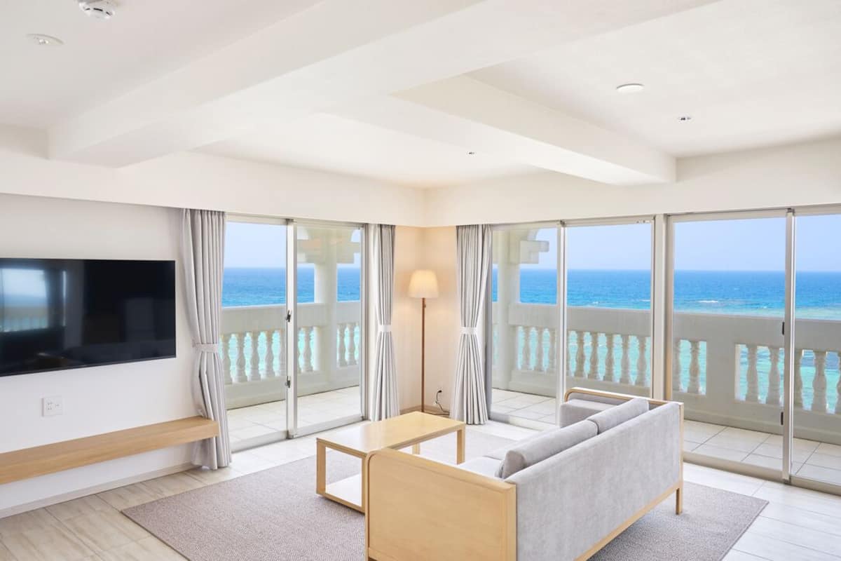 Habitación de hotel moderna con un televisor grande, un sofá, una mesita y un balcón con vistas al océano.