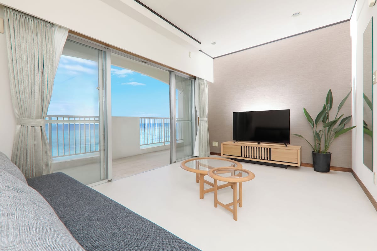 A modern living room with a large TV, a sofa, two glass tables, and a balcony with a view of the sea.