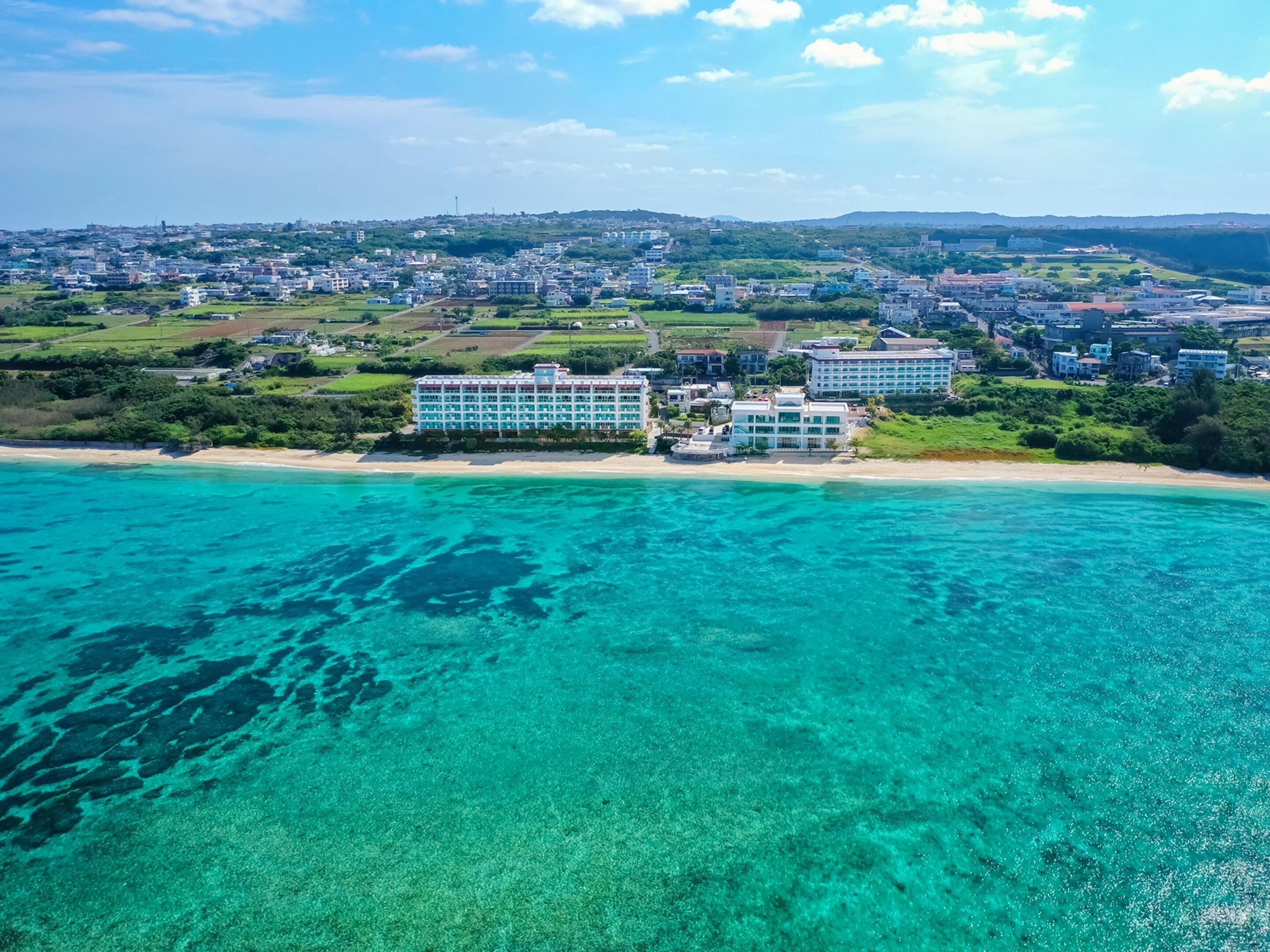Ein Küstenhotel mit mehreren Gebäuden, einem Strand und klarem türkisblauem Wasser.