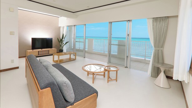 Een moderne woonkamer met een bank, televisie en glazen tafels, met uitzicht op een balkon en de zee.
