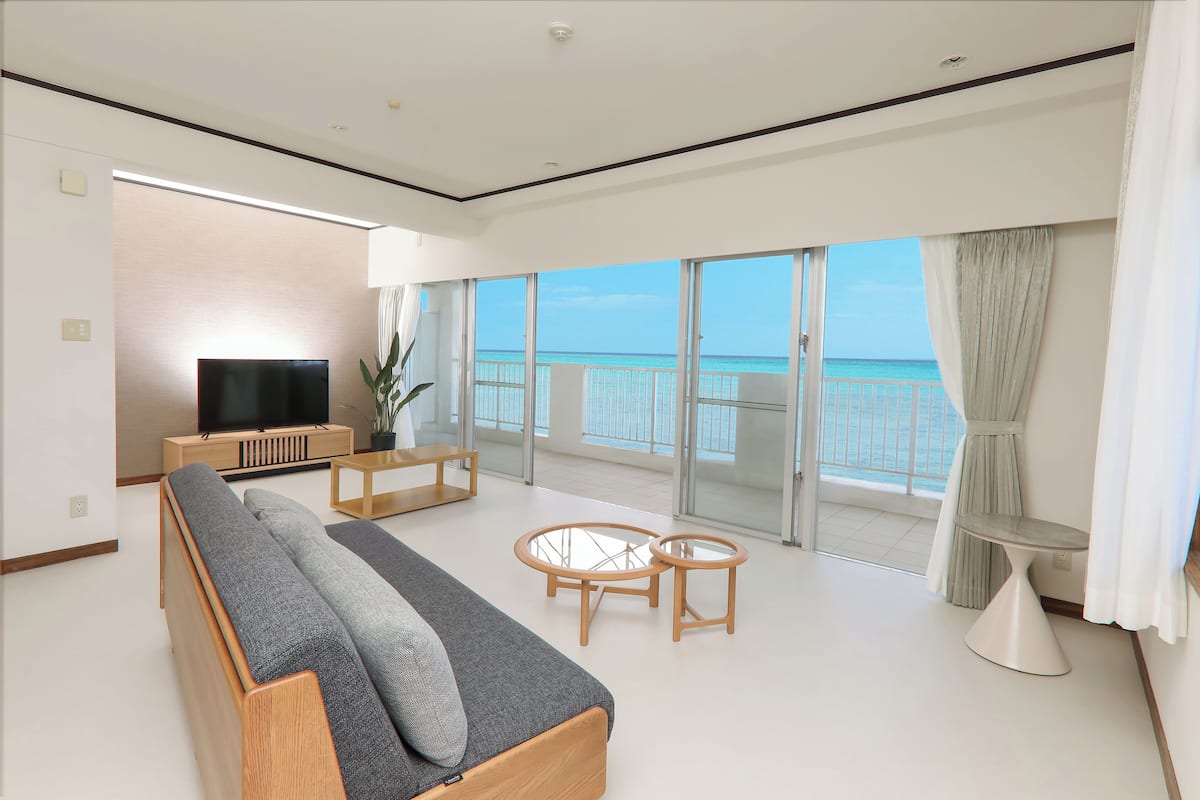 A modern living room with a sofa, TV, and glass tables, overlooking a balcony with a view of the sea.