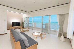 Una sala de estar moderna con sofá, televisor y mesas de vidrio, con vistas a un balcón y al mar.