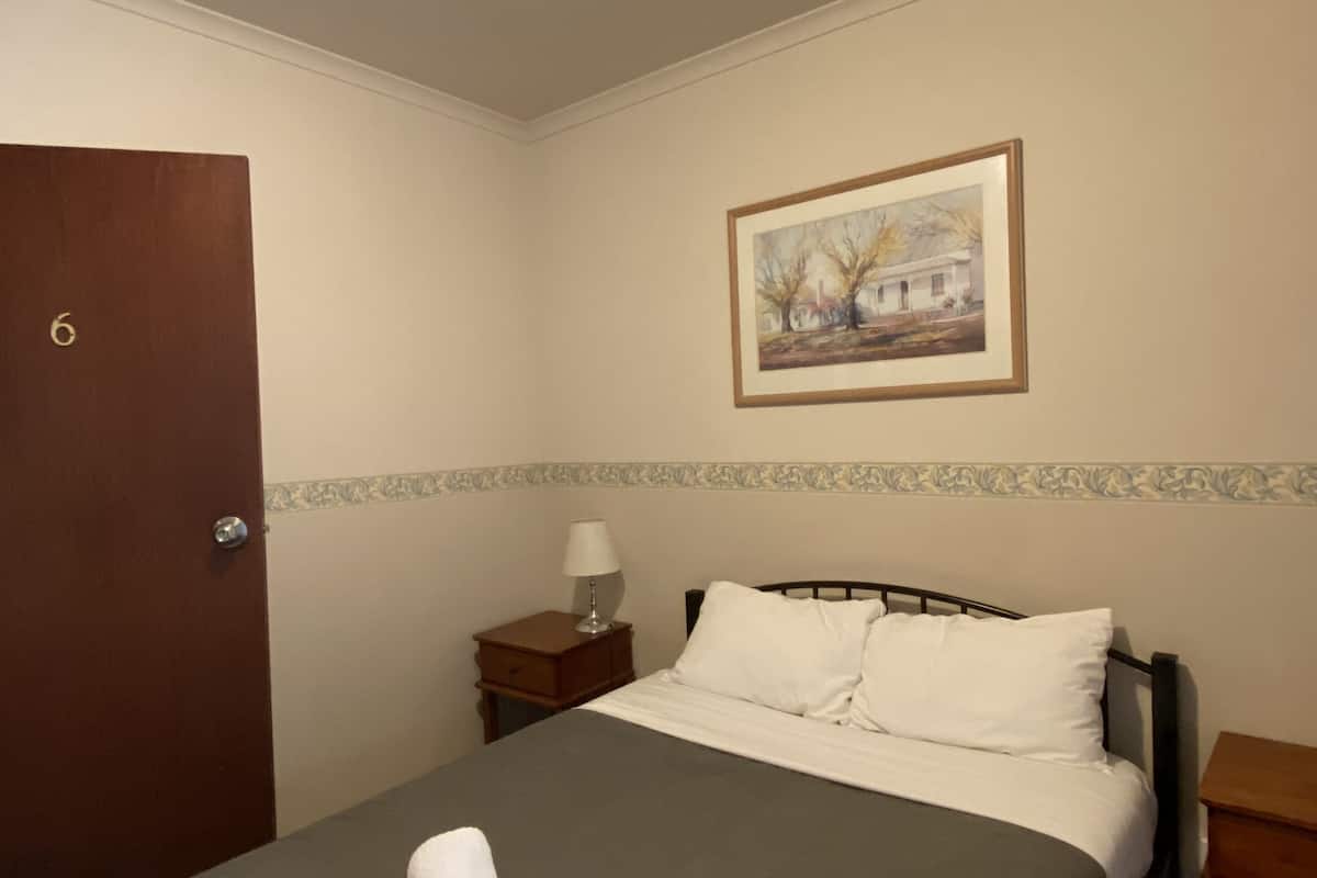 Un lit bien fait, avec du linge de lit blanc et deux serviettes enroulées, une table de chevet avec une lampe, un tableau encadré au mur et une porte avec un numéro.