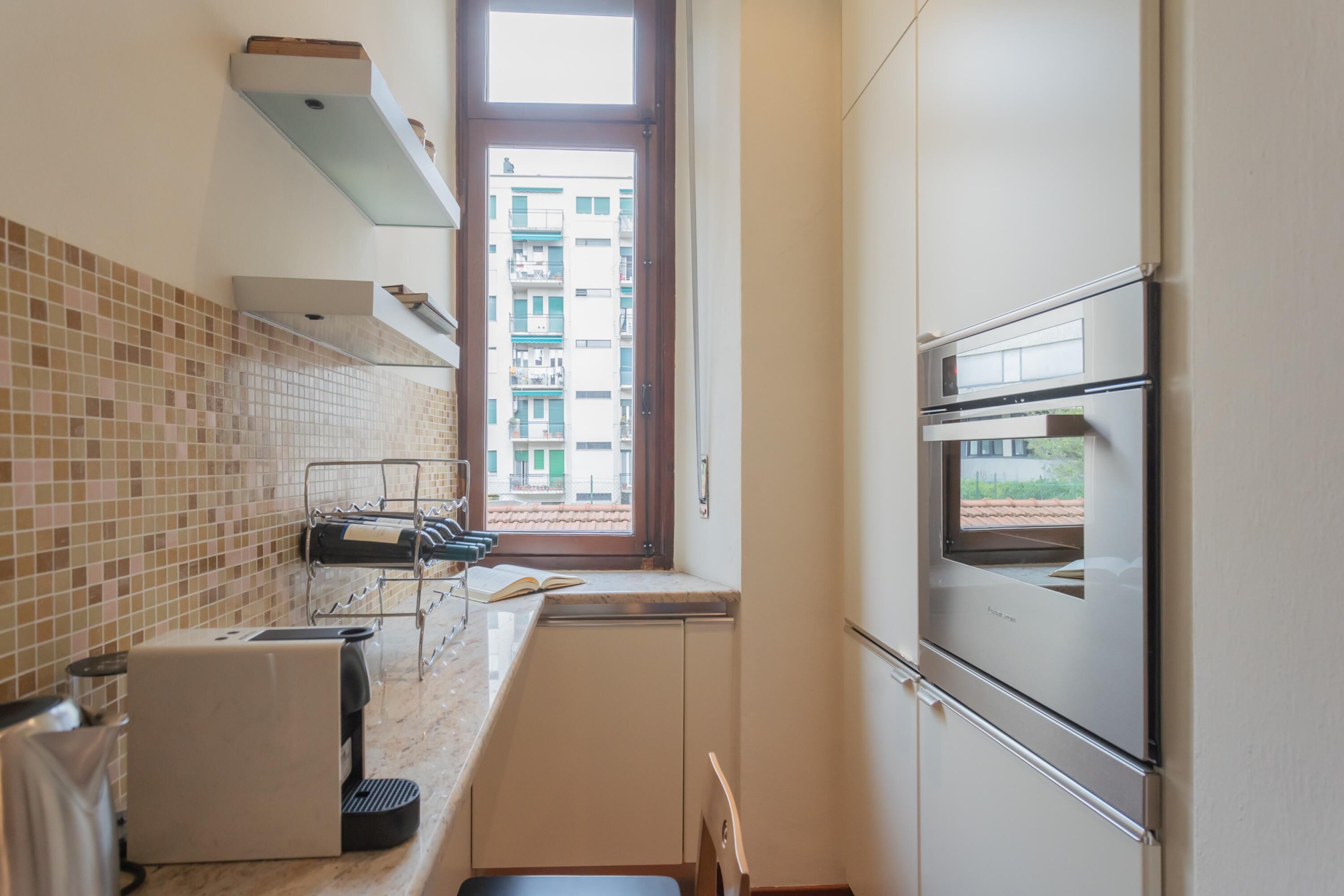 A compact kitchen with a built-in oven, a coffee machine, and a window with a view of a building.