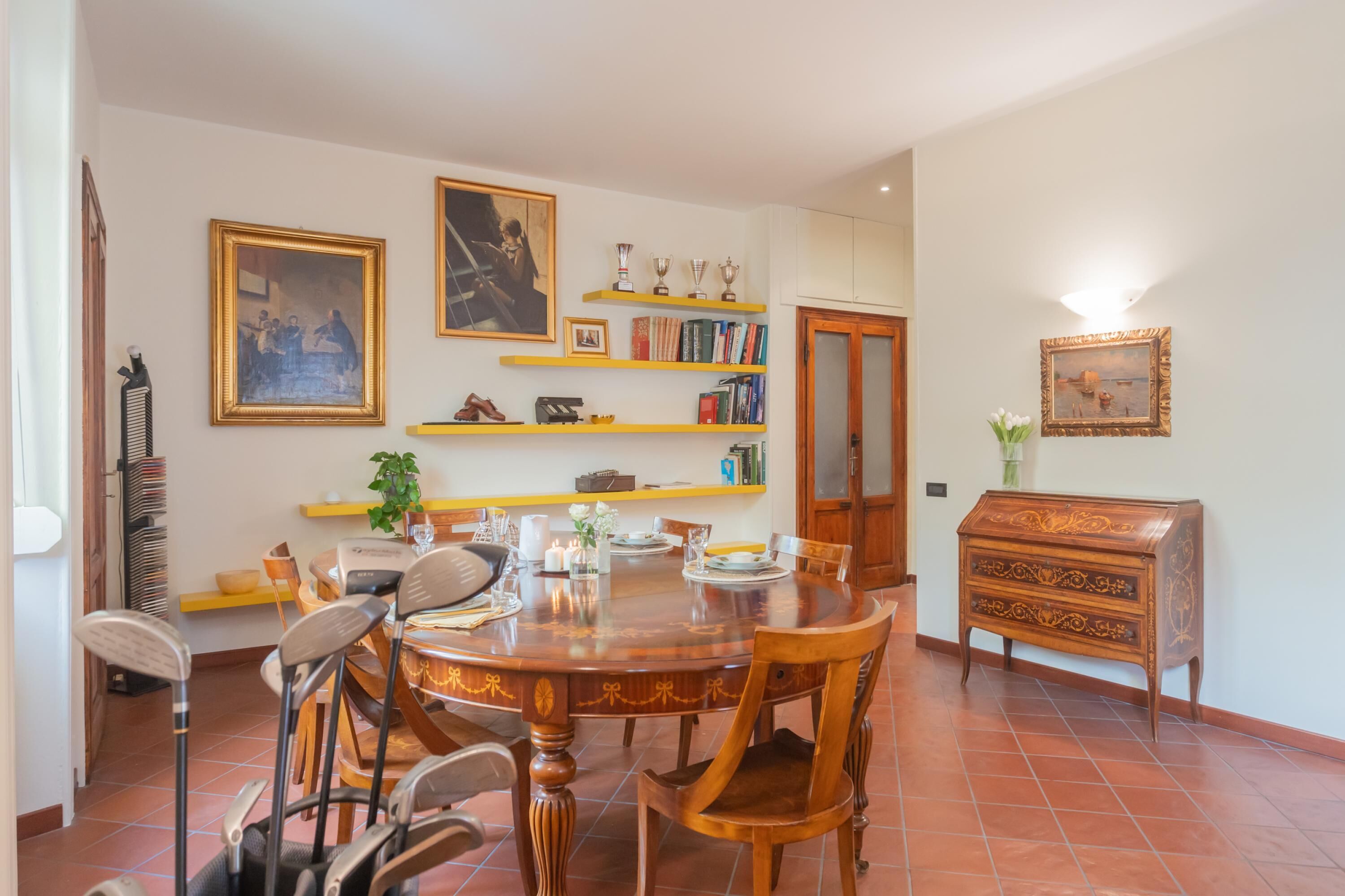 A dining area with a round wooden table, chairs, and a bookshelf with books and trophies.