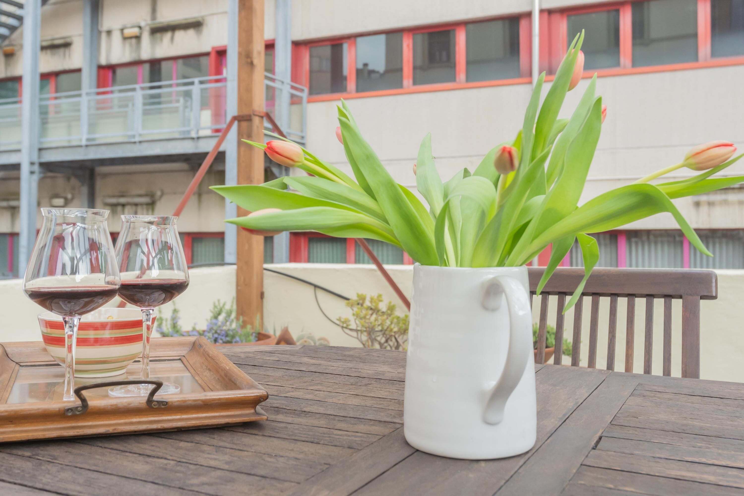 A wooden table with a white pitcher and tulips, and two glasses of red wine on a tray.