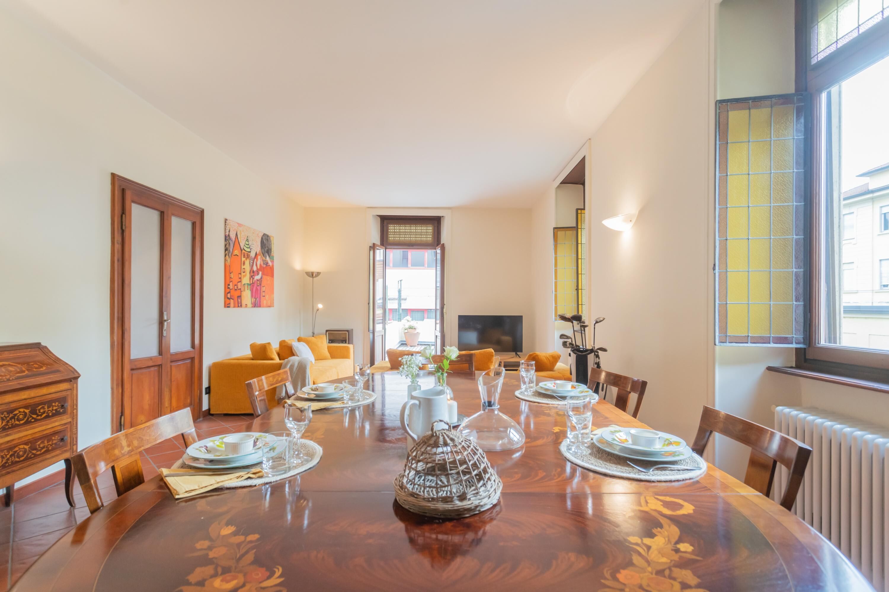 A dining area with a wooden table set for four, surrounded by chairs. There is a sofa and a television in the background.