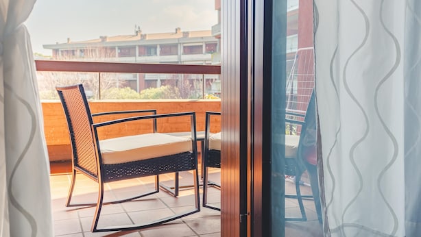 A balcony with a chair and a table, a view of buildings in the background, and a curtain partially drawn. - Fior di Loto - Italian Homing (Desenzano del Garda)