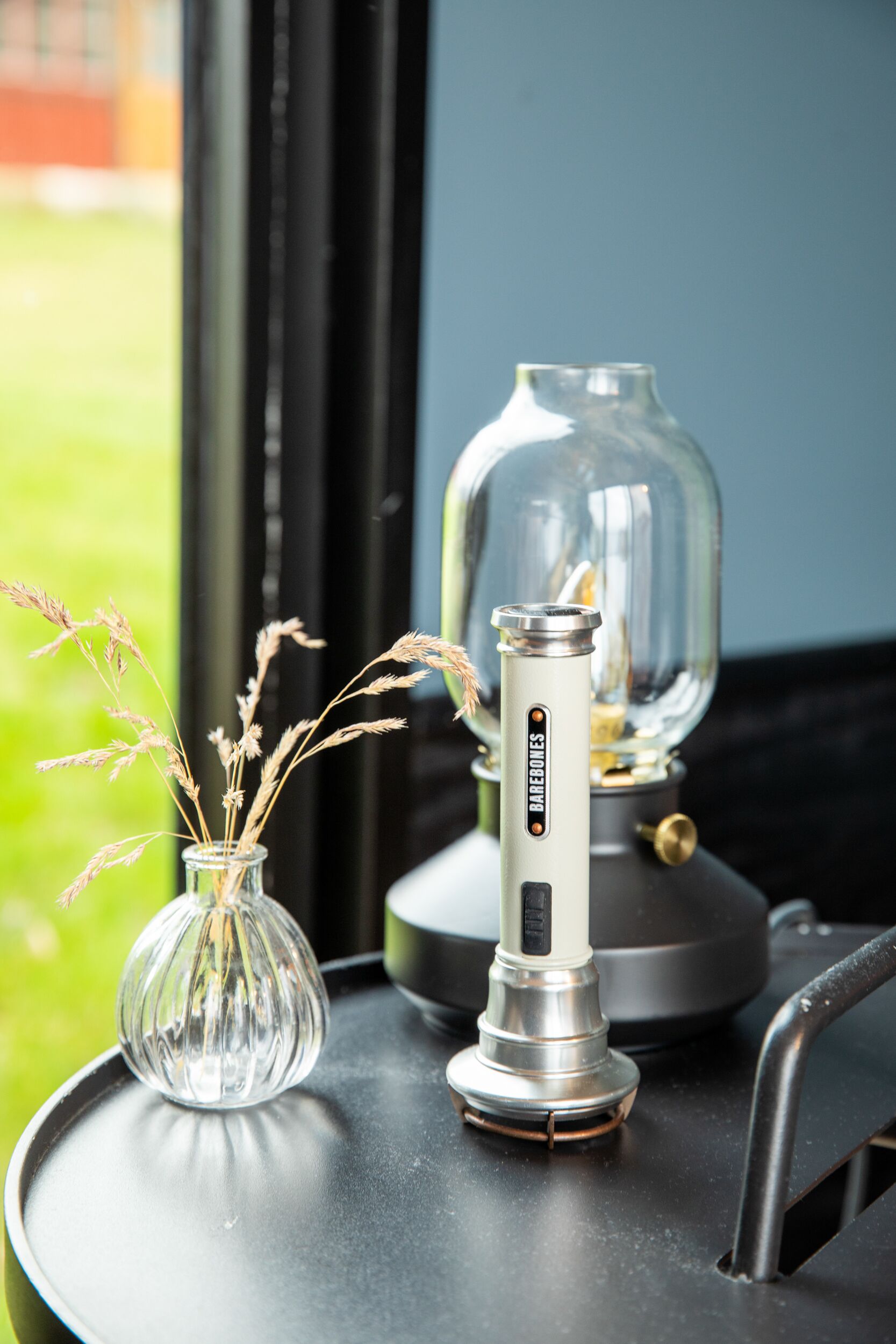 Una mesa con un florero de vidrio transparente que contiene plantas secas y un dispositivo eléctrico moderno con acabado metálico.