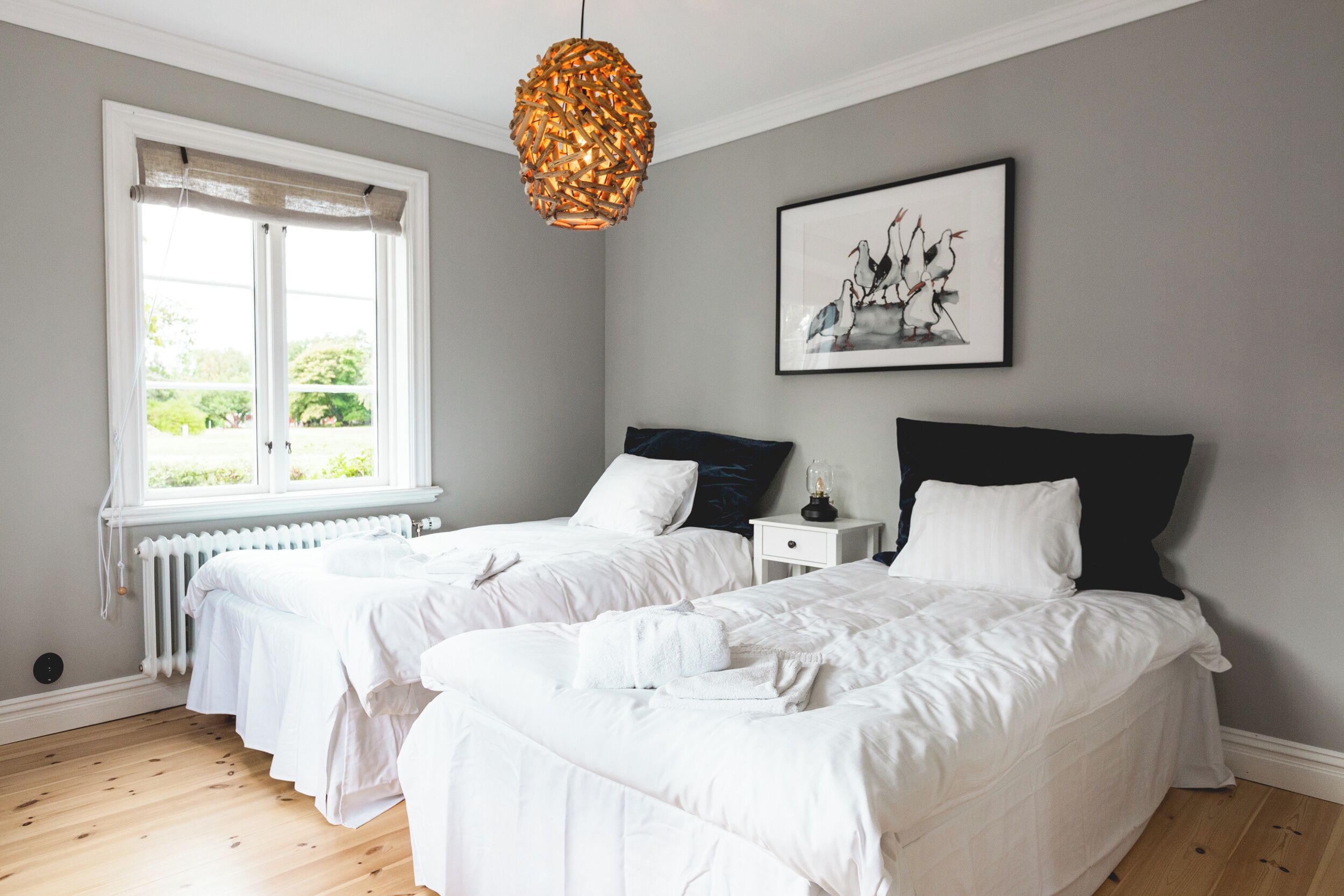 A hotel room with two single beds, a window with a view of greenery, a radiator, and a hanging light fixture.