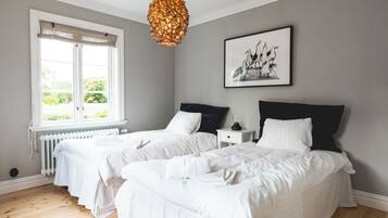 A hotel room with two single beds, a window with a view of greenery, a radiator, and a hanging light fixture.