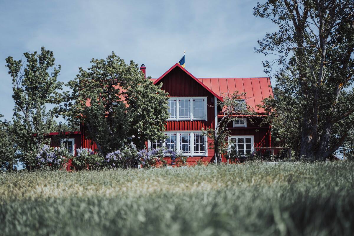 A red two-story house with a red roof, surrounded by trees and purple flowers.