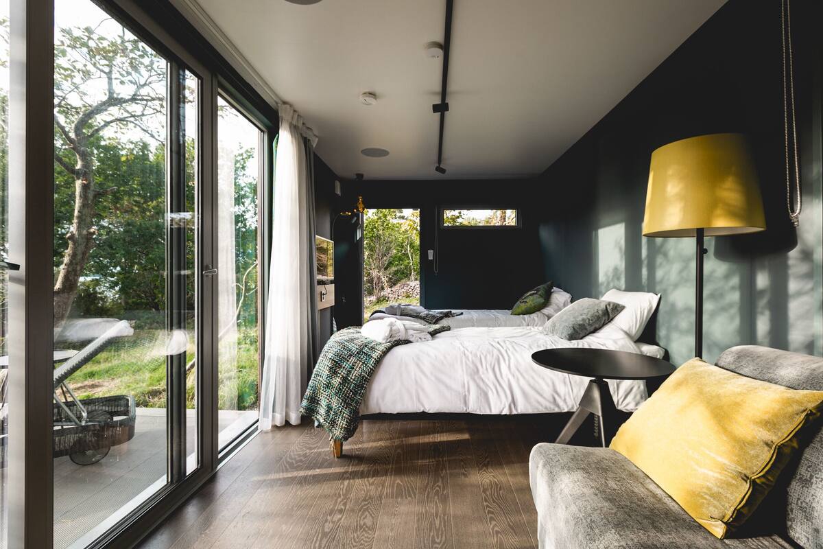 A bedroom with a large bed, a bedside table, a chair, and a window with a view of greenery.