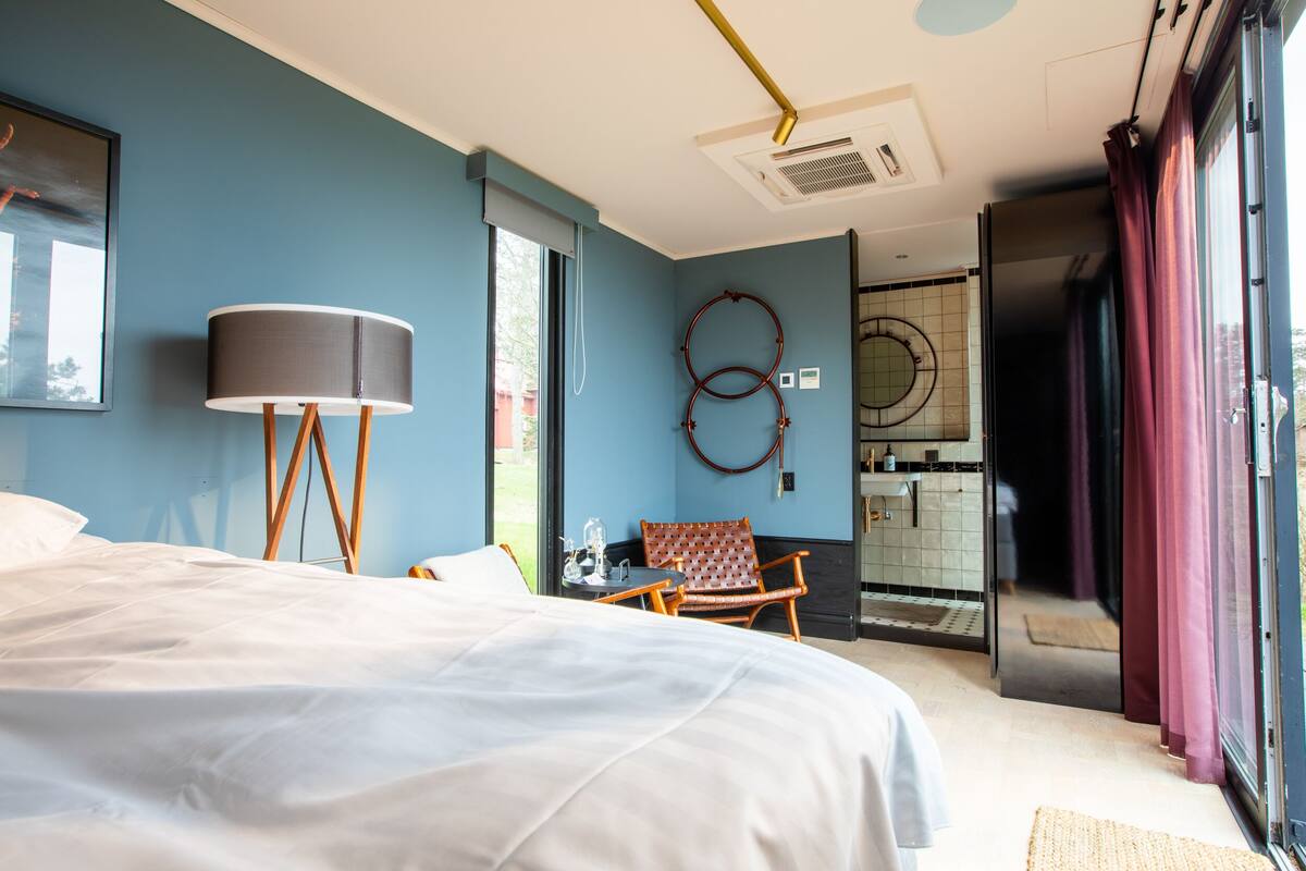 A modern hotel room with a large bed, a desk, a chair, and a view of the bathroom and outside.