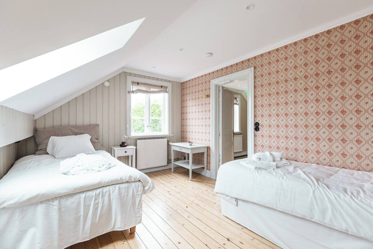A bedroom with two beds, a small table, a radiator, and a window with curtains.