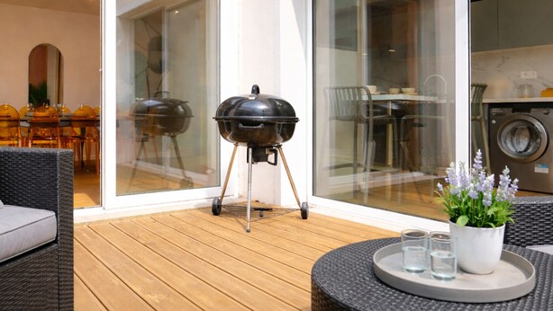A patio with a barbecue grill, a round table with glasses and a plant, and a view of a dining area through glass doors.