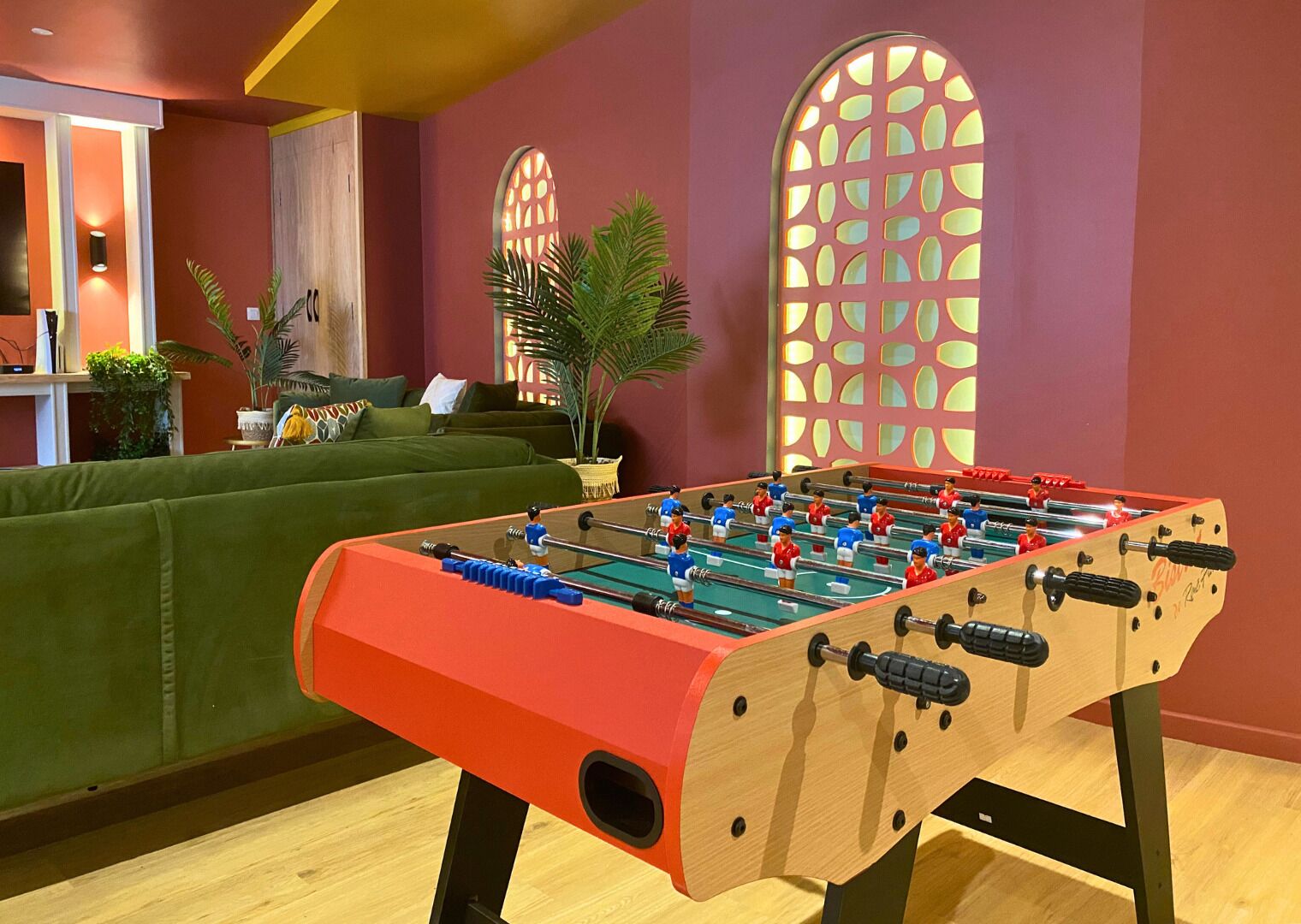 A foosball table in a room with a green sofa, two windows with decorative grills, and a plant.