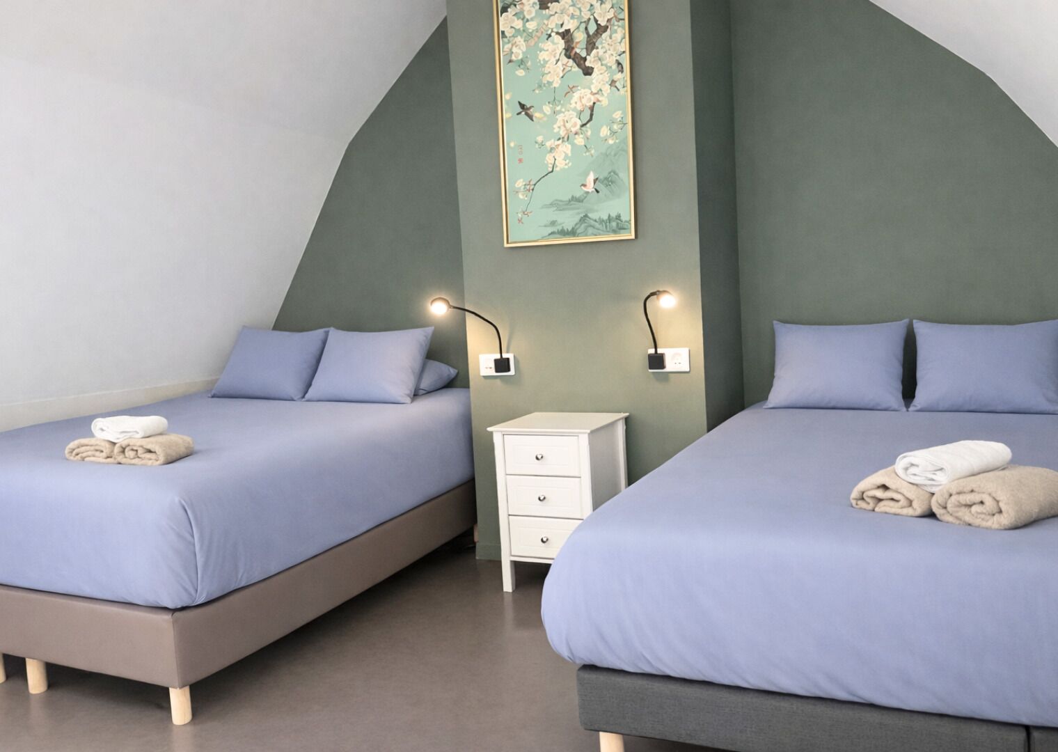 A hotel room with two beds, each with a pillow and a rolled towel. There is a nightstand with a lamp between the beds.