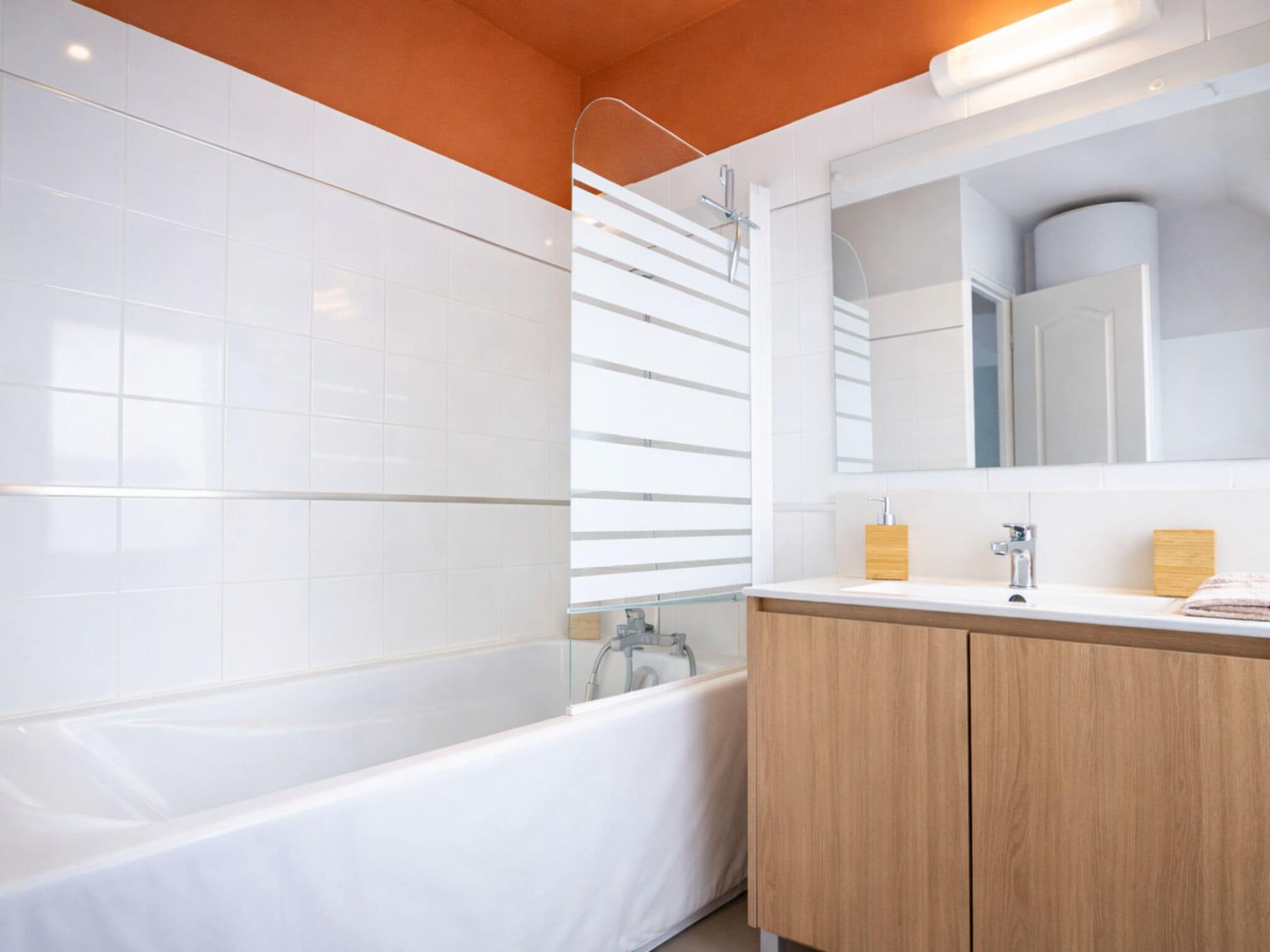 A modern bathroom with white tiles, a bathtub, a shower with a glass door, and wooden cabinets.