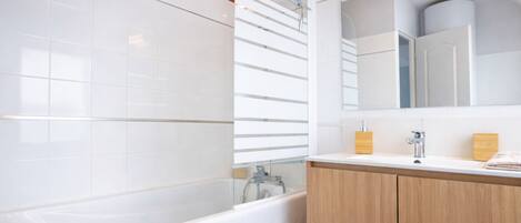 A modern bathroom with white tiles, a bathtub, a shower with a glass door, and wooden cabinets.