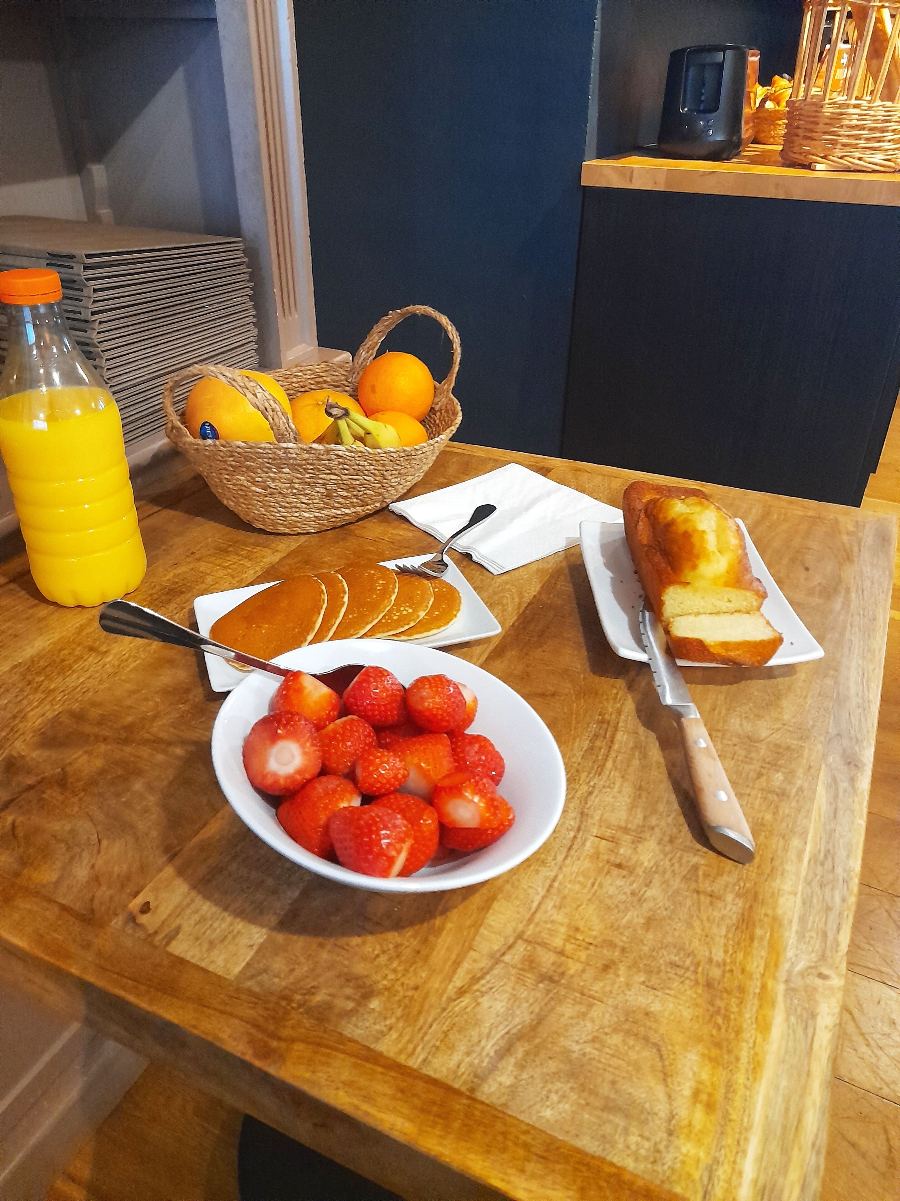 A breakfast table with orange juice, strawberries, pancakes, a loaf of bread, and a basket of oranges.