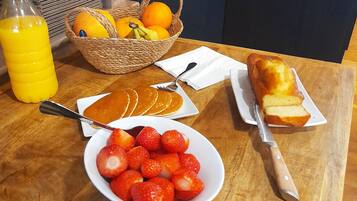 A breakfast table with orange juice, strawberries, pancakes, a loaf of bread, and a basket of oranges.