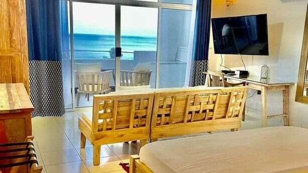 A hotel room with a large bed, a desk with a TV, and a balcony with a view of the ocean. - Beau Vallon Studios (Mahé Island)