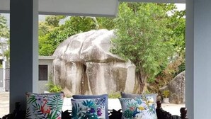 A covered seating area with a sofa and cushions, a wooden table, and a view of a large rock and trees. - Beau Vallon Studios (Mahé Island)