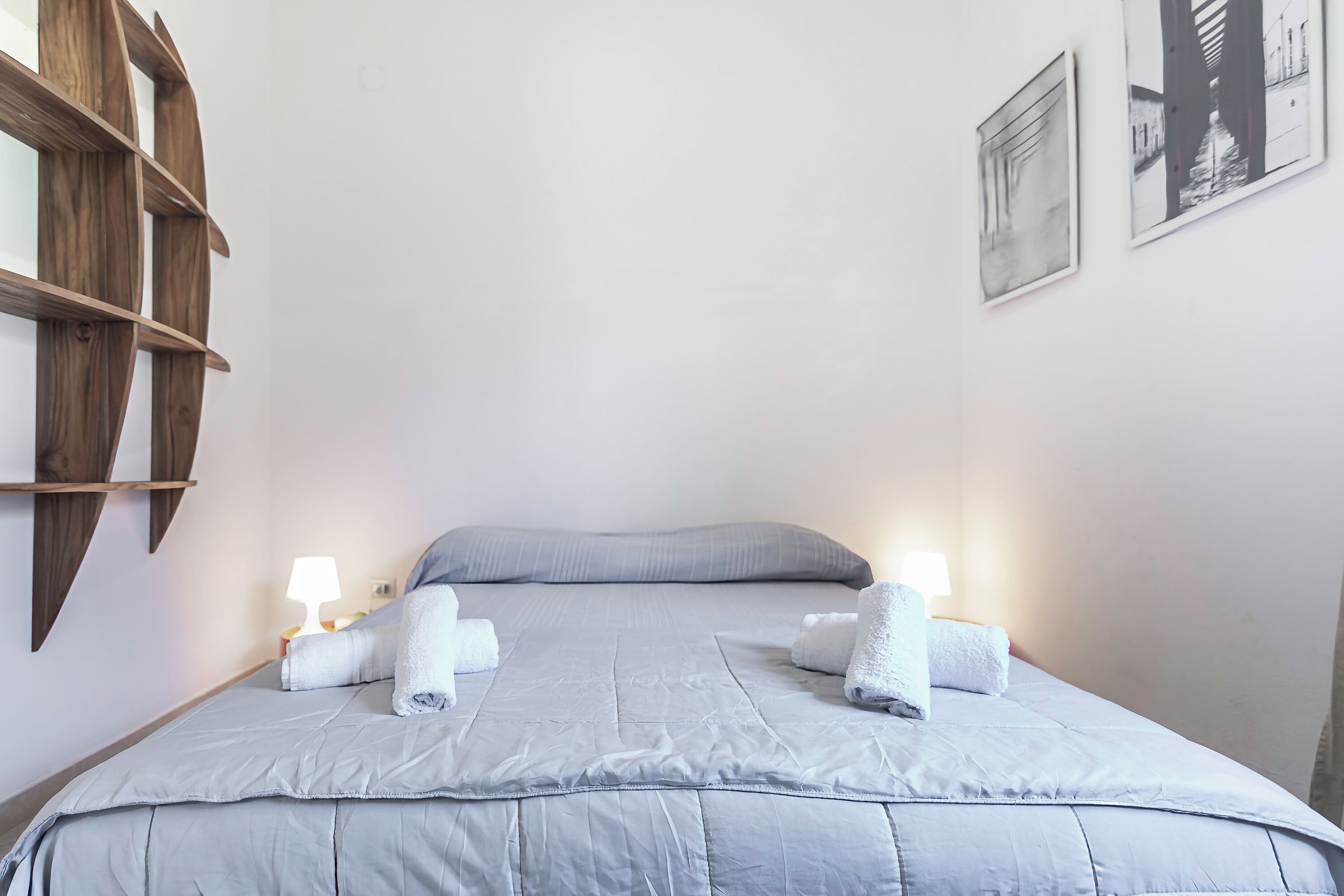 A neatly made bed with a grey duvet, two pillows, and two lit bedside lamps.