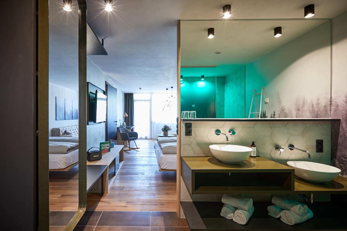 Una habitación de hotel moderna con un espejo grande, dos camas, una zona de estar y un baño con un lavabo y espejo amplios.