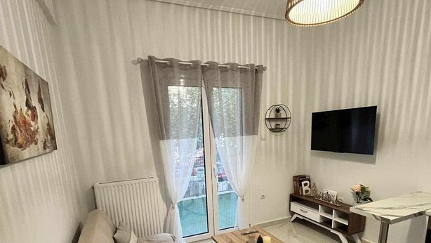 En moderne stue med en beige sofa, et træbord, et fjernsyn ophængt på væggen og et stort vindue med gardiner.