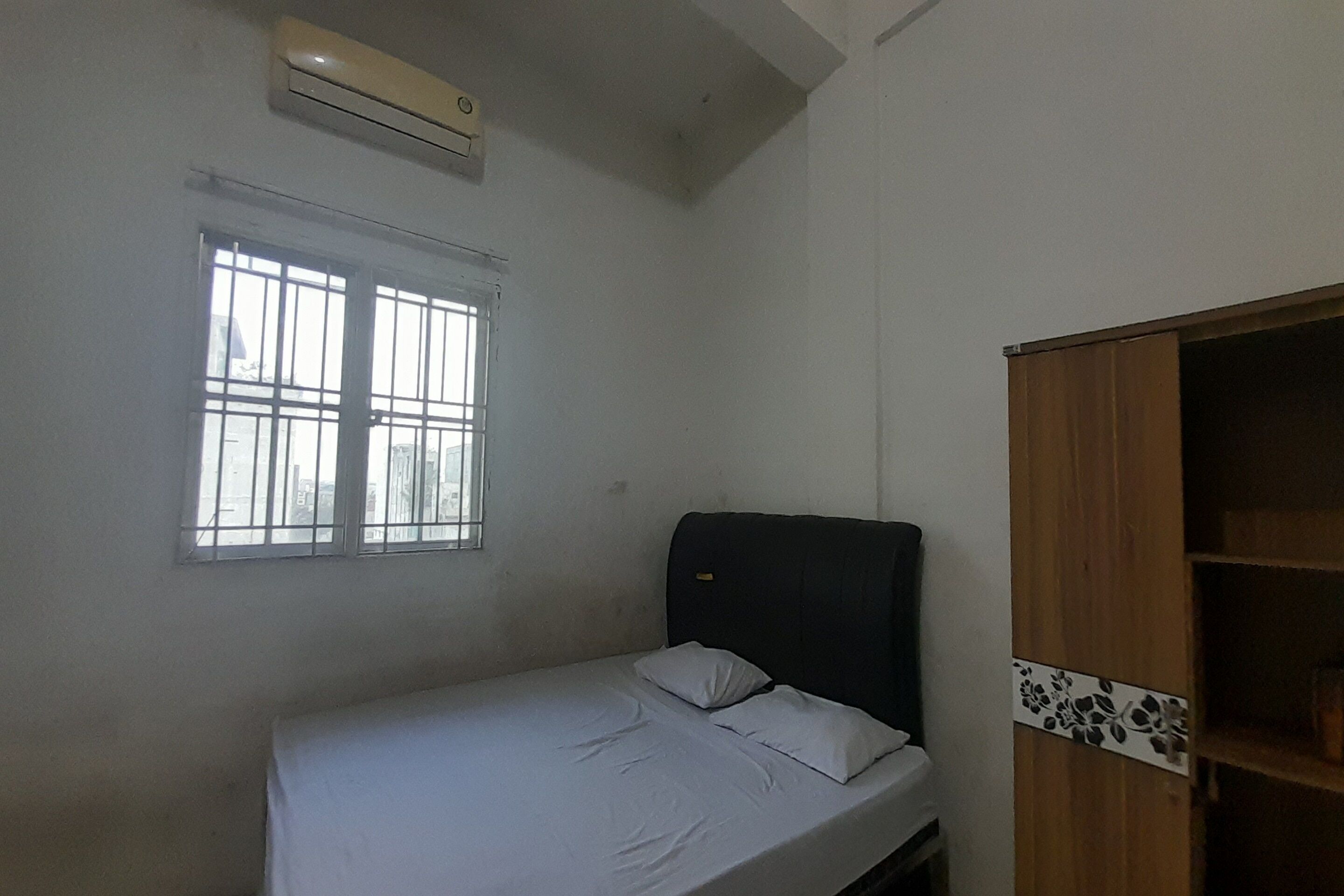 Une petite chambre avec un lit simple, une armoire en bois, une fenêtre avec des barreaux et un appareil de climatisation fixé au mur.