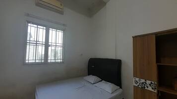 Une petite chambre avec un lit simple, une armoire en bois, une fenêtre avec des barreaux et un appareil de climatisation fixé au mur.