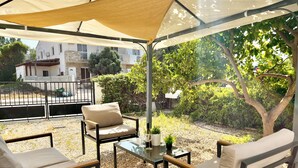 Un patio al aire libre con una zona de estar techada, una mesa de vidrio y vistas a un edificio residencial y vegetación.