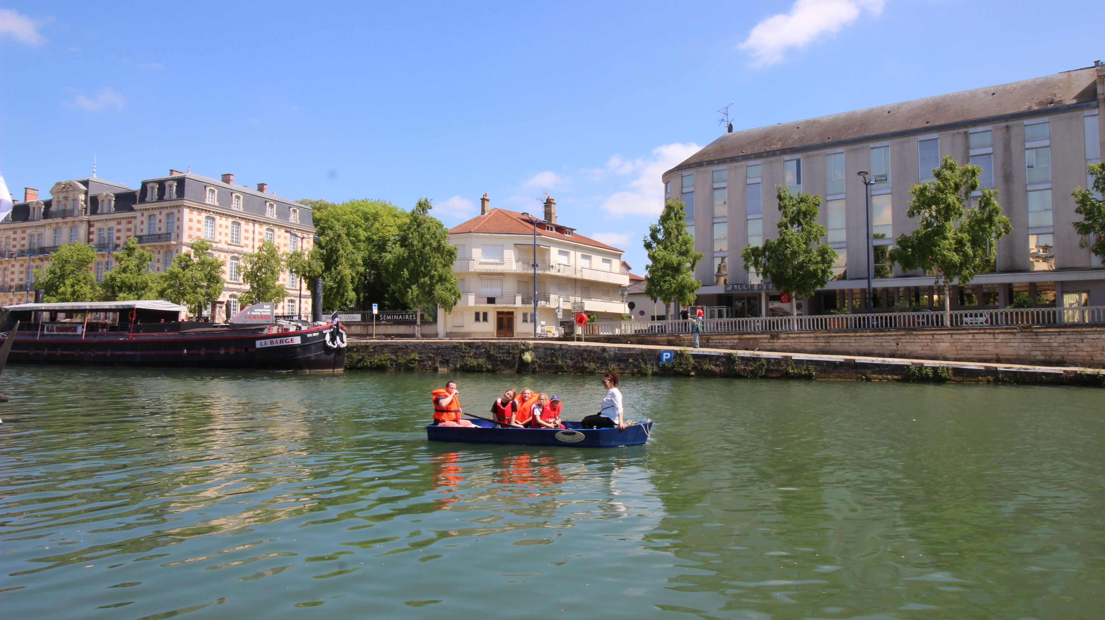 A river with a boat carrying passengers, buildings along the riverbank, and a bridge.