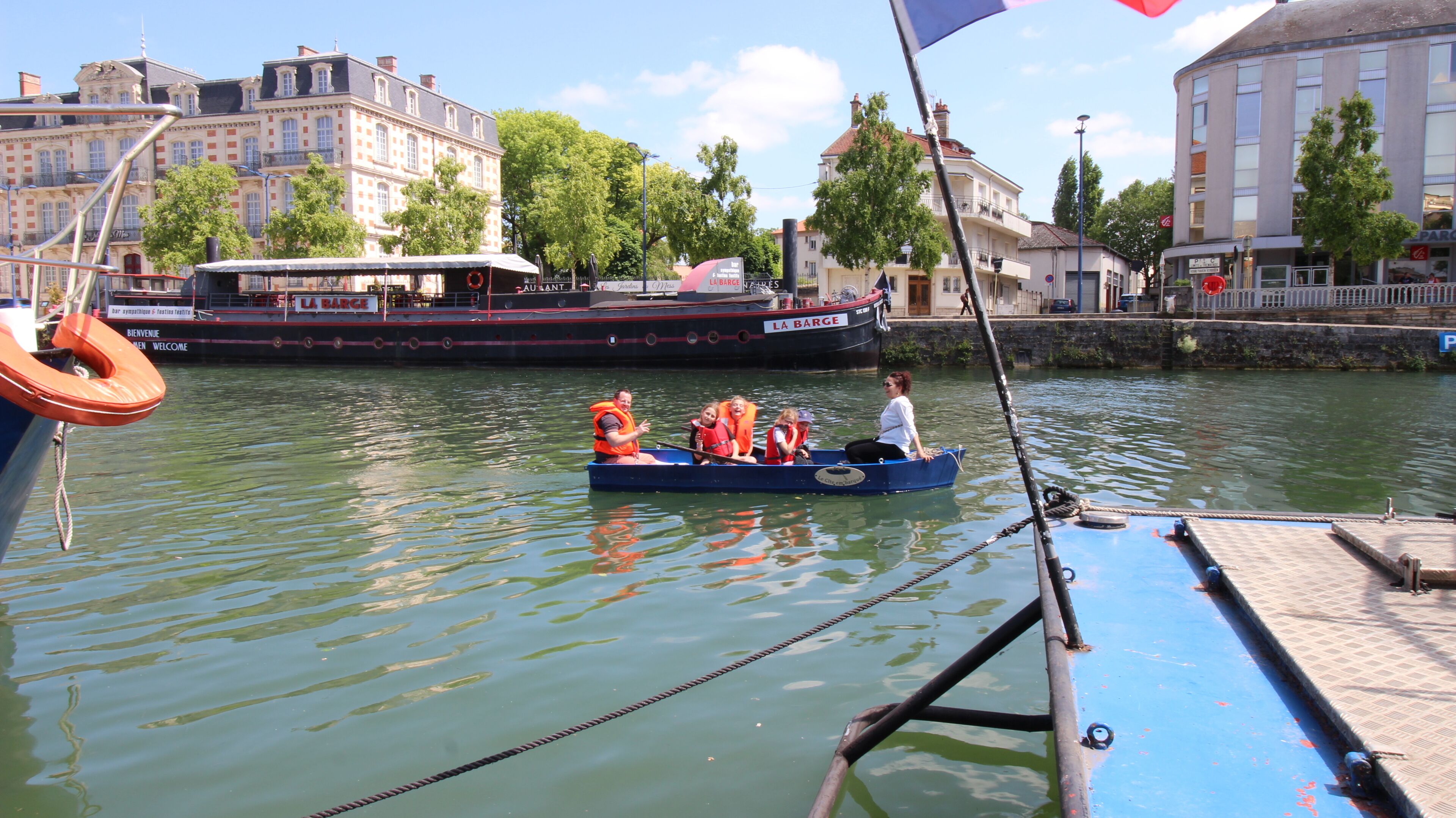 Ein Boot mit Passagieren in Rettungswesten, vertäut neben einem Gebäude mit der Aufschrift 'LA BARGE'.