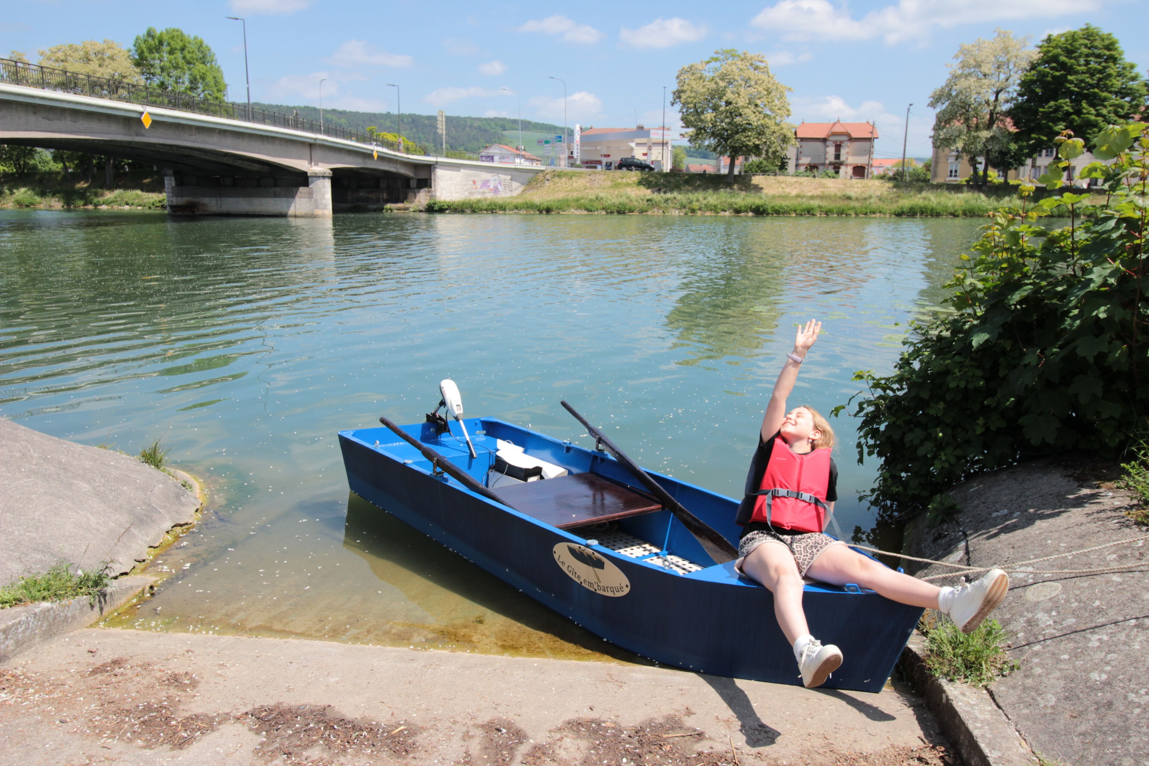 A river with a bridge, a blue boat, and a person in a red life jacket.