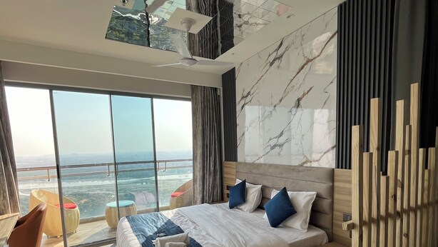 A modern hotel room with a large bed, a view of the city, and a balcony with wicker furniture. - Luxsugero Studios (Noida)