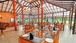 Uma ampla sala de jantar com mesas e cadeiras de madeira, teto alto com vigas de metal e grandes janelas que permitem entrada de luz natural.