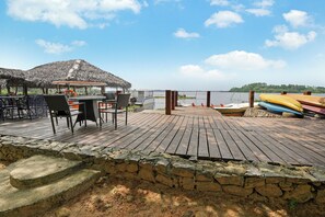 A wooden deck with a thatched-roof structure, a table with chairs, and a view of the water.
