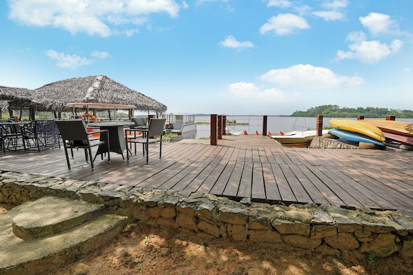 Una terraza de madera con una estructura de techo de paja, una mesa con sillas y vista al agua.